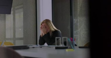 Kadın, modern ofiste masa başında otururken yüzünü iki eliyle kapatıyor. Sakin bir şirket ortamında stres ya da duygusal hayal kırıklığını ifade ediyor.