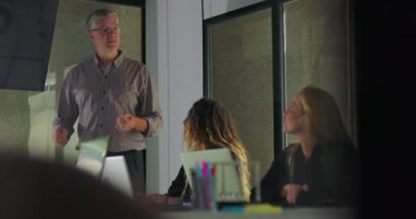 Kadın meslektaşlar dijital ekrandaki veri tablolarına bakarken, erkek profesyoneller konferans odasında grup sunumu yapıyor.