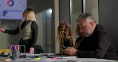 İş adamı ofis toplantısı sırasında gizlice telefonu kontrol ediyor. Kadın meslektaşları sunum yapıyor. Gündelik modern iş yeri dinamikleri ve çoklu görev ve sessiz takım çatışması.