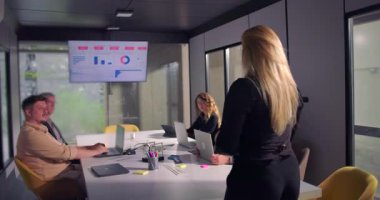 Takım, olağan ofis toplantısı sırasında modern konferans odasında toplandı. Kadın meslektaşlar dikilip verileri sunarken, iş ortamında odaklanma ve tartışma sağlandı.