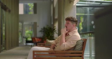 Ofisteki bankta oturan bir adam, elleri çenesinde, düşünceli ve düşünceli görünürken sessiz çalışma alanındaki uzaklığa bakıyor.