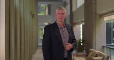 Orta yaşlı bir adam modern ofis koridorunda duruyor ceketini ayarlıyor, kendine güvenen ve parlak bir profesyonel ortamda yaşıyor.