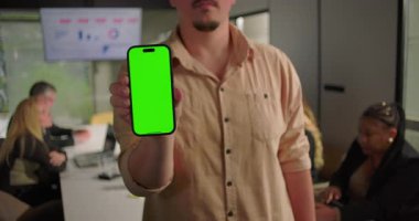 Ofiste yeşil ekranlı akıllı telefonu tutan genç adam, konferans masasında arka planda meslektaşlarıyla kameraya doğru kolunu uzatıyor.