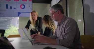 İş kadını, modern konferans odasında ekranda veri tablolarıyla iş arkadaşlarıyla birlikte bilgisayarı incelemek için masaya yaslanıyor.