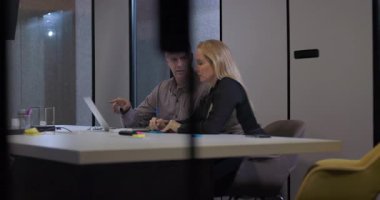 Konferans masasında tartışan erkek ve kadın iş arkadaşları laptoptaki bilgileri gözden geçiriyorlar, modern iş yerinde geç saatlerde iş birliği yapıyorlar.