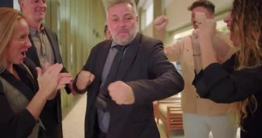 Çeşitli iş adamları yüksek enerjili iş yeri kutlamaları sırasında coşkuyla tezahürat yapıp alkışlarken, ofis koridorunda coşkulu erkek çalışanlar çılgınca kutlama yapıyor.