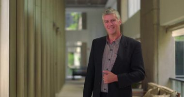 Modern ofis koridorunda gülümseyen orta yaşlı bir adam, kendine güvenen ve parlak profesyonel çalışma alanında ulaşılabilir görünüyor.