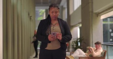 Ofis koridorunda yürürken akıllı telefon kullanan Afro-Amerikalı kadın, arka planda oturan iş arkadaşları ile ekrana odaklandı