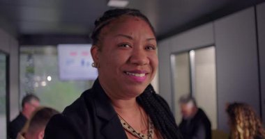 Gülümseyen Afrikalı Amerikalı iş kadını portresi, özgüvenli ve dostane bir ifade, arka planda çalışan çeşitli takımlar.