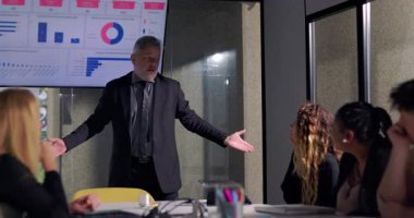 Kızgın işletme yöneticisi, meslektaşları ekranda gösterilen çizelgelerle kritik geri bildirimleri dinlerken toplantı odasında gergin bir toplantı sırasında ekibe işaret ediyor.