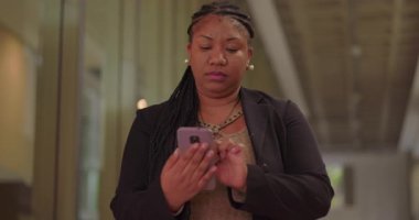 Koridorda dikilirken akıllı telefon kullanan odaklanmış Afro-Amerikan kadın ofis çalışma alanındaki yansıtıcı cam duvar ve tavan detaylarıyla