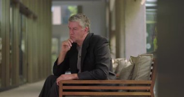 Orta yaşlı bir adam ofiste sessizce oturuyor, elleri çenesinde, huzurlu modern çalışma alanında düşünceli ve düşünceli görünüyor.