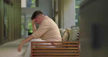 Ofis koridorunda elleri kapalı bir şekilde bankta oturan adam, sakin bir mesleki ortamda hayal kırıklığını ya da zihinsel yorgunluğu ifade ediyor.