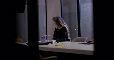 Stresli genç iş kadını toplantı masasında tek başına oturuyor. Şirket toplantısından sonra, ofis malzemeleri ve dijital cihazlarla çevrili.