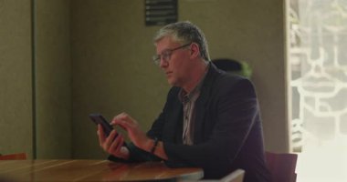 Ofis odasındaki ahşap masada otururken akıllı telefon kullanan gri saçlı ve gözlüklü olgun bir adam profesyonel ortamdaki cihaza odaklanmış.