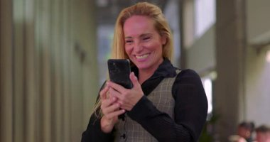 İş kadını telefon kamerasındaki dişleri parlak bir gülümsemeyle kontrol ediyor. Modern ofis koridorunda dururken ruj izi kalmadığından emin oluyor.