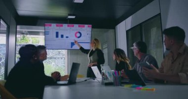 İş kadını harita ekranının yanındaki ekibe seslenirken, meslektaşlar da parlak ışıklandırılmış konferans odasında şirket toplantısını izliyorlar.