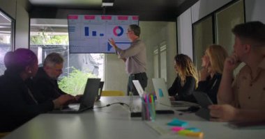 Erkek yönetici modern konferans odasında çeşitli iş ekiplerine veri sunarken, ekranda tablolar ve meslektaşlar toplantı masasının etrafında dinliyorlar.