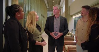 Şirket yöneticisi modern ofis koridorunda çeşitli takımlara sesleniyor, bir grup profesyonel gayri resmi toplantı sırasında iş yerinde sohbet ediyorlar.