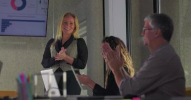 İş kadını, sunumdan sonra açık bir jest yaparak gülümserken, meslektaşları kurumsal ofiste takım toplantısını alkışlıyor.