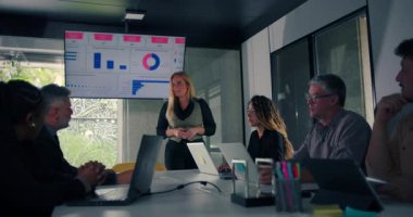 Sarışın iş kadını, modern ofisteki veri ekranıyla konferans masasının etrafında oturan nişanlı meslektaşlarına sunum yapıyor.