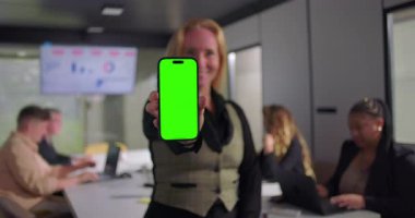 İş kadını akıllı telefon ekranına gülümseyerek yeşil ekran cihazını kameraya doğru genişletiyor. Konferans masasında çalışan çeşitli takımlar ve monitörde veri grafikleri var.