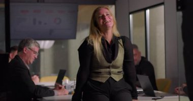 Üst düzey iş kadını, modern ofis yönetim kurulundaki toplantı sırasında dizüstü bilgisayarlarla çalışan meslektaşlarıyla birlikte konferans masasında otururken güvenle gülümsüyor.