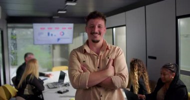 Ofiste kollarını kavuşturmuş gülümseyen genç adam, olumlu ve kendinden emin ifade, konferans masasında ekran verileriyle uğraşan meslektaşlar