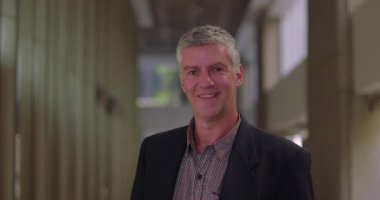 Modern ofis koridorunda duran gülümseyen orta yaşlı işadamı, doğrudan kameraya bakıyor, işyeri koridorunda kendine güvenen profesyonel bir erkek.