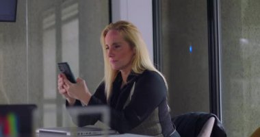 Modern konferans odasında teknoloji ve konsantrasyonla ofis masasında otururken akıllı telefon ekranına odaklanmış bir iş kadını.