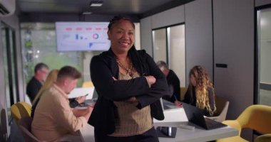 Gülümseyen Afrikalı Amerikalı iş kadını, kollarını kavuşturmuş vaziyette duruyor. Ekip olarak özgüvenli liderlik mevcudiyeti, veri ekranında işbirliği yapıyor.