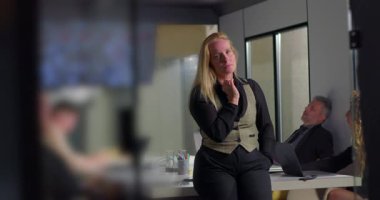 Ofis toplantısı sırasında konferans masasına yaslanan sarışın iş kadını, ekranda arka planda bulanık analizleri gösteriyor