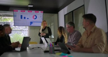 Sarışın iş kadını dijital tabloya işaret ederken cam duvarlı konferans odasındaki toplantı sırasında nişanlı ekibe istatistikleri açıklıyor