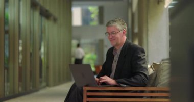 Orta yaşlı bir adam rahat ofis ortamında dizüstü bilgisayarla çalışırken hafifçe gülümsüyor, modern koridor çalışma alanında rahatça oturuyor.