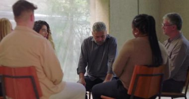 Adam grup terapisi sırasında konuşur, öne doğru eğilir ve sessiz odada diğerleriyle doğal ışık ve duygusal odaklanma dolu destek çemberine girer.