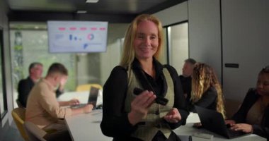 Gülümseyen iş kadını, gördüklerinden hoşlandıktan sonra yeşil ekranlı yatay akıllı telefonu ofis arka planında çalışan çeşitli takımlara doğru uzattı.