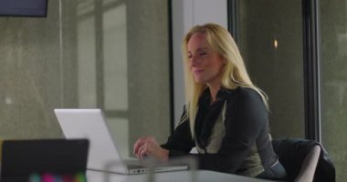 Modern ofis toplantı odasında dizüstü bilgisayarın başında oturmuş kameraya bakan gülümseyen iş kadını, iş günü boyunca sıcaklık ve dostluk saçıyor.