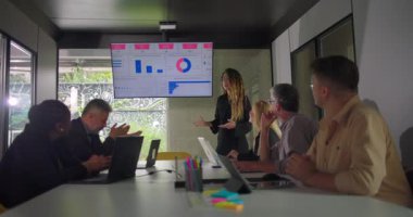 Genç sunucu veri tablosuna işaret ederken, takım modern ofiste dizüstü bilgisayarlar ve duvara monte edilmiş ekranla toplantı sırasında dinliyor