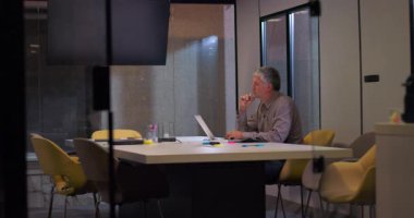 Profesyonel erkek, akşam ışığında ofis masasında tek başına çalışır. Odaklanmış ifadeyle, belgelerin ve renkli ofis malzemelerinin arasında dizüstü bilgisayarda yazar.