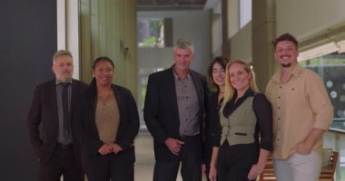 Altı kişilik çok kültürlü bir iş grubu koridorda duruyor, gülümseyen profesyoneller kapalı alanda doğal ışık alan çalışma alanındaki kameralara bakıyorlar.