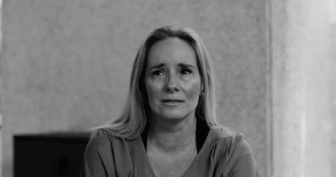 Kadın doğrudan kameraya bakıyor gözyaşları içinde derin üzüntü ve kırılganlık ifade ederken sessiz, samimi bir şekilde içeride oturuyor, siyah ve beyaz.