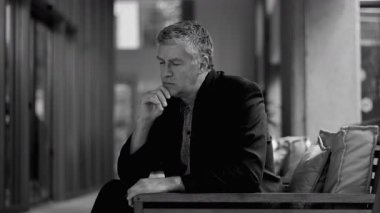 İş kıyafetleri içinde yaşlı bir adam gözleri kapalı ve elleri çenesinde otururken modern iç mekanda ciddi düşüncelere dalmış.