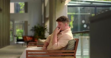 Ofis salonundaki bankta oturan bir adam, parlak modern çalışma alanında biraz rahatsız ya da utangaç görünür.