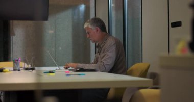 Kıdemli iş adamı ofis konferans masasında tek başına oturur dizüstü bilgisayarla çalışır, sakin bir akşam iş ortamında görevine odaklanır.