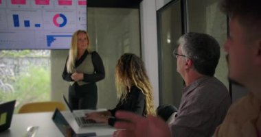 Ekranın önünde duran iş kadını, modern ofis ortamında masanın etrafında oturan meşgul ekibe verileri açıklıyor.