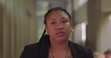 Örgü ören Afro-Amerikalı kadın ciddi görünüyor ve arka planda dikey paneller ve doğal ışık ile modern ofis koridorlarında bestelenmiş.