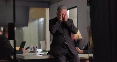 İş adamı zorlu ofis toplantılarında yüzünü iki eliyle kapatırken, meslektaşları da şirket yönetim kurulunun arka planında dizüstü bilgisayarlar üzerinde çalışıyor.