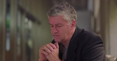 Orta yaşlı bir adam, kederli bir ifadeyle mesafeye bakıyor. Ofis salonunda oturuyor ve iç kargaşa anında elleri önlerine kenetlenmiş.