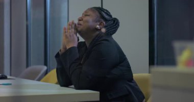 Afrikalı Amerikalı iş kadını, elleri birbirine bastırılmış bir şekilde ofis masasında tek başına oturuyor, duygusal olarak Tanrı 'ya dua ediyor ve kişisel kriz zamanlarında güç için yalvarıyor.