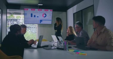 Modern ofisteki iş toplantısında masadaki bilgisayarları kullanarak çeşitli meslektaşlarıyla duvar ekranında verileri sunan genç kadın dikkatle dinliyorlar.